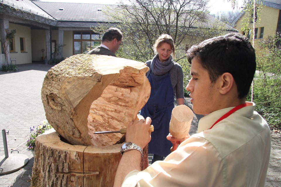 Das Bild zeigt einen mann der mit einem Schnitzeisen und Hammer ein Stück Holz bearbeitet.