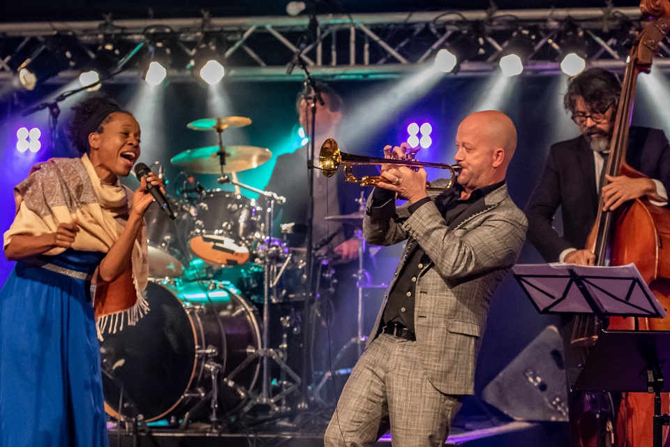 Das Bild zeigt eine Sängerin, einen Trompeter, einen Bassisten und einen Schlagzeuger auf der Bühne.