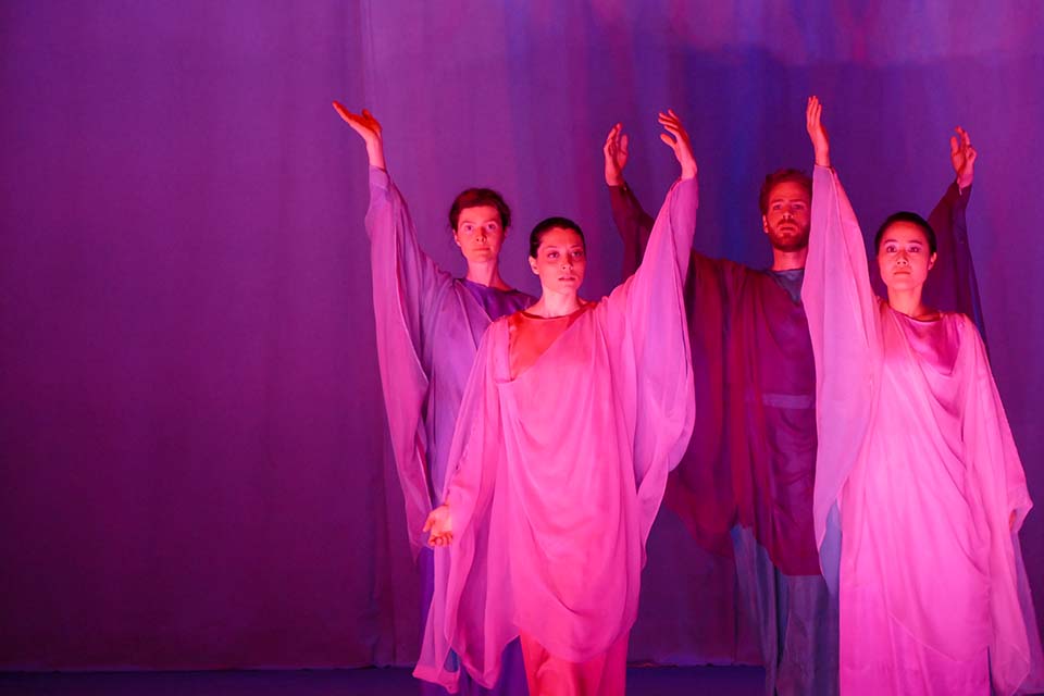 Das Bild zeigt drei Frauen und einen Mann in rosa-violett farbenen Gewändern auf einer Bühne.